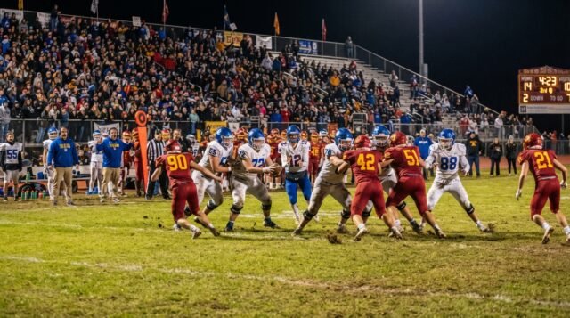 High School Football Game Under Friday Night Lights High school football players competing under Friday night stadium lights in front of packed bleachers in the United States.